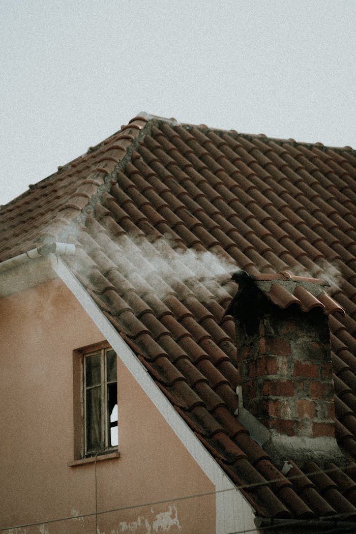 A house with a tiled roof releasing smoke from a chimney, indicative of heating or cooking activities.